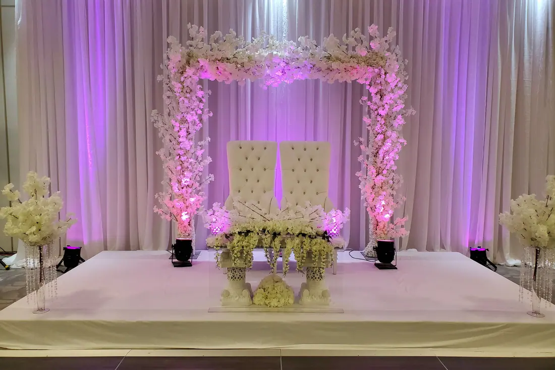 Raised wedding stage with an arch of white cherry blossom over twin tufted chairs against full-height ivory draping