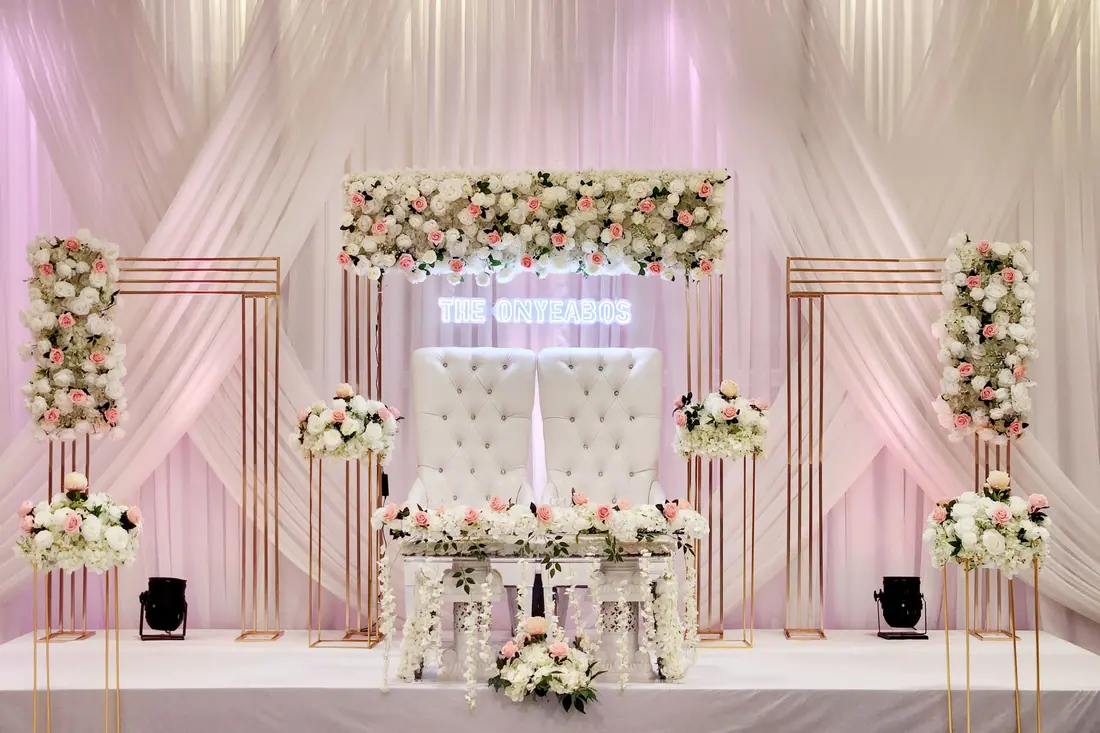 Sweetheart table framed by blush and ivory floral arrangements on slim gold stands with a custom neon sign above