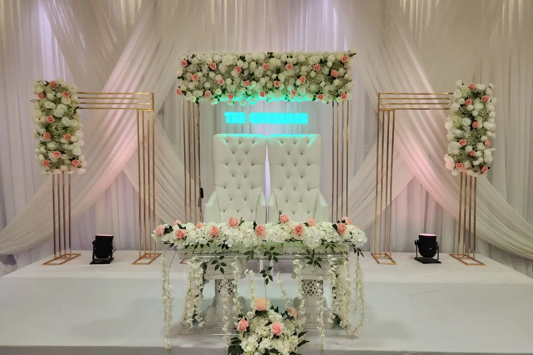 Blush and white floral wedding backdrop with a neon name sign, gold frames and a mirrored head table with trailing greenery