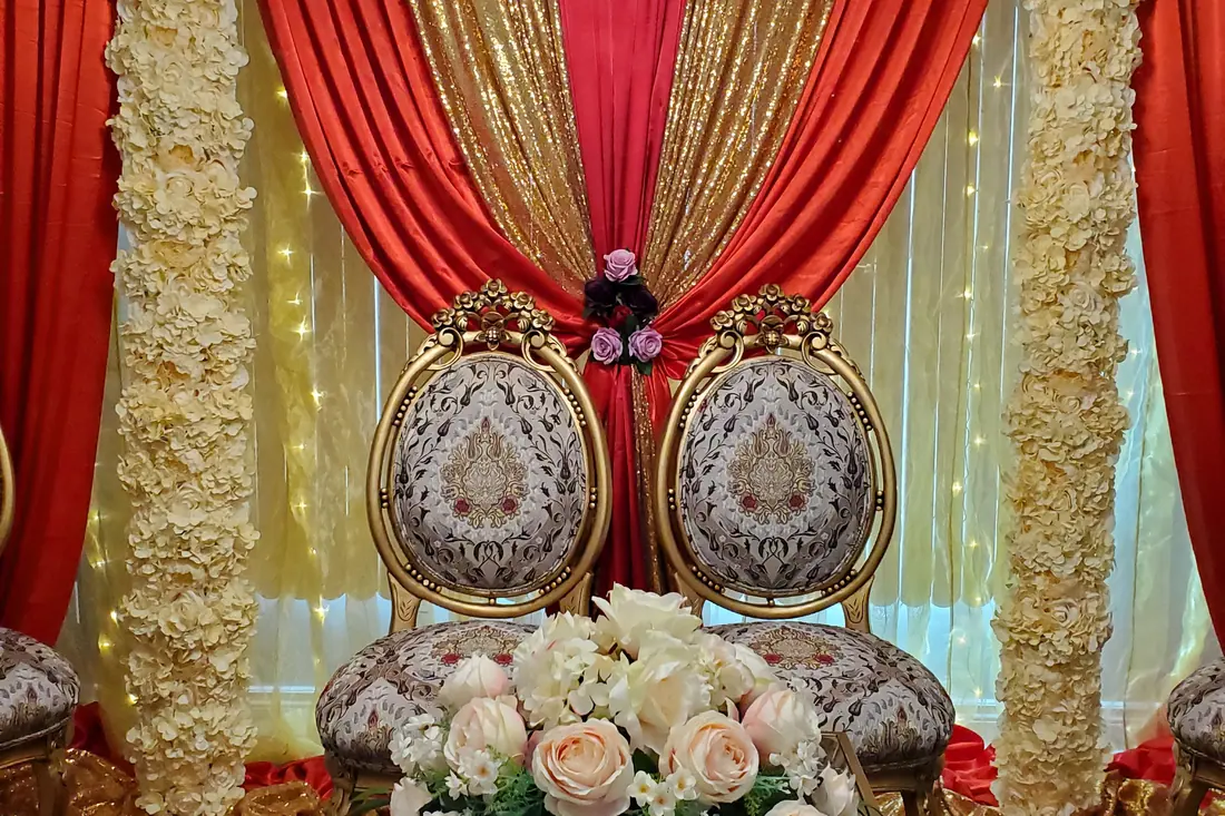 Ornate carved gold and ivory wedding chairs in front of red and gold draping with rose-covered wall panels and fairy lights