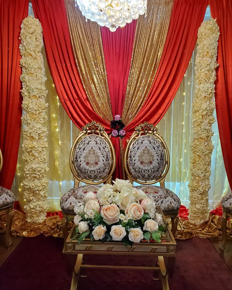 Ornate carved gold and ivory wedding chairs in front of red and gold draping with rose-covered wall panels and fairy lights