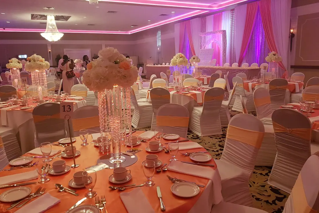 Reception hall of white covered chairs with coral sashes and tall floral pomander centerpieces under warm coral uplighting