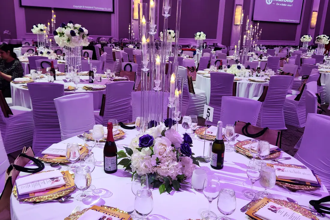 Rows of tall crystal candelabras and white floral arrangements on long tables in a violet-lit event hall