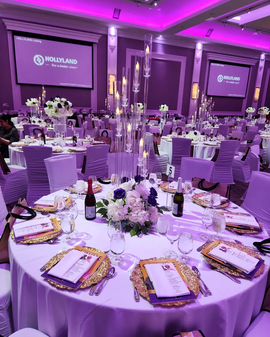 Rows of tall crystal candelabras and white floral arrangements on long tables in a violet-lit event hall