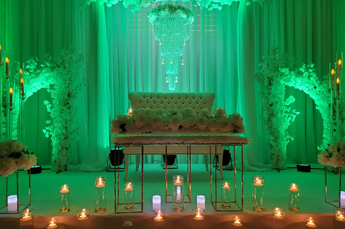Wedding stage in emerald green uplighting with floral archways, a tufted bench and a row of gold candle holders