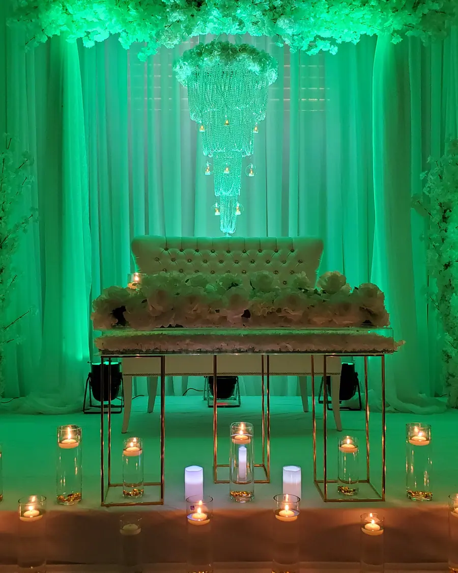 Wedding stage in emerald green uplighting with floral archways, a tufted bench and a row of gold candle holders