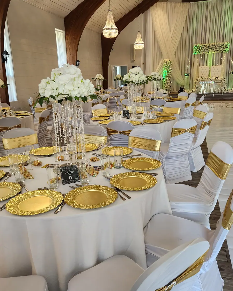 Bright ivory wedding tablescape with gold charger plates, gold chair sashes and a tall crystal centerpiece topped with white hydrangea