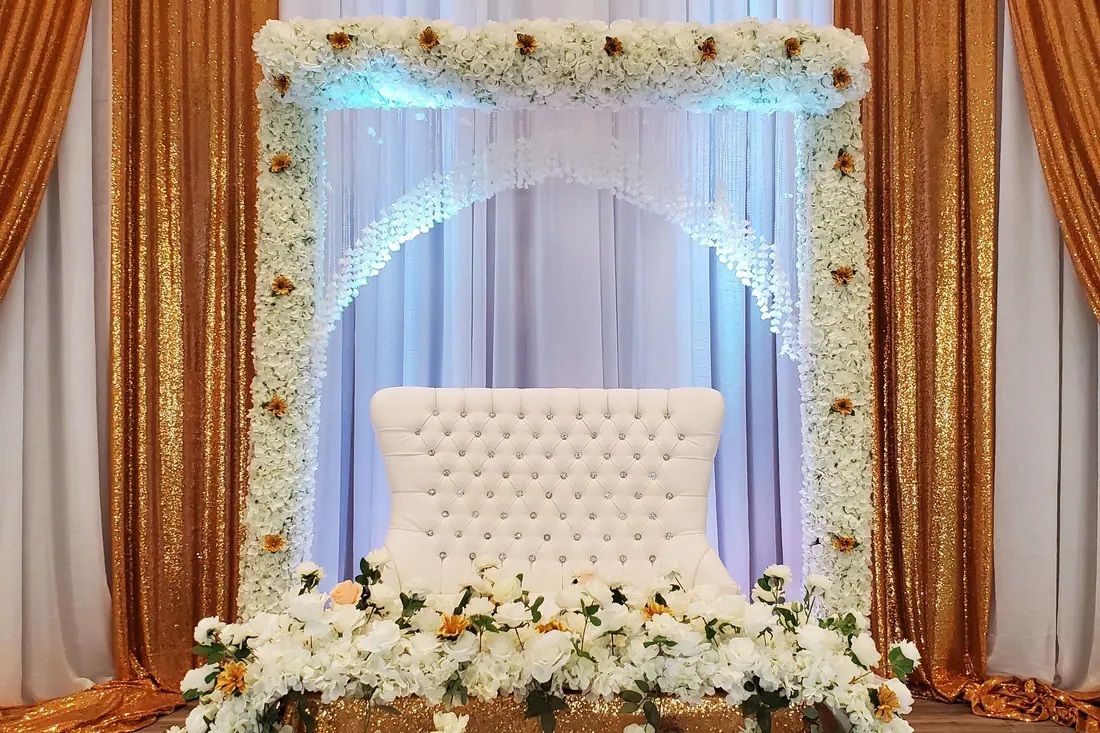 Gold draped wedding backdrop with a white floral arch and teal uplighting behind a tufted bench and low white florals