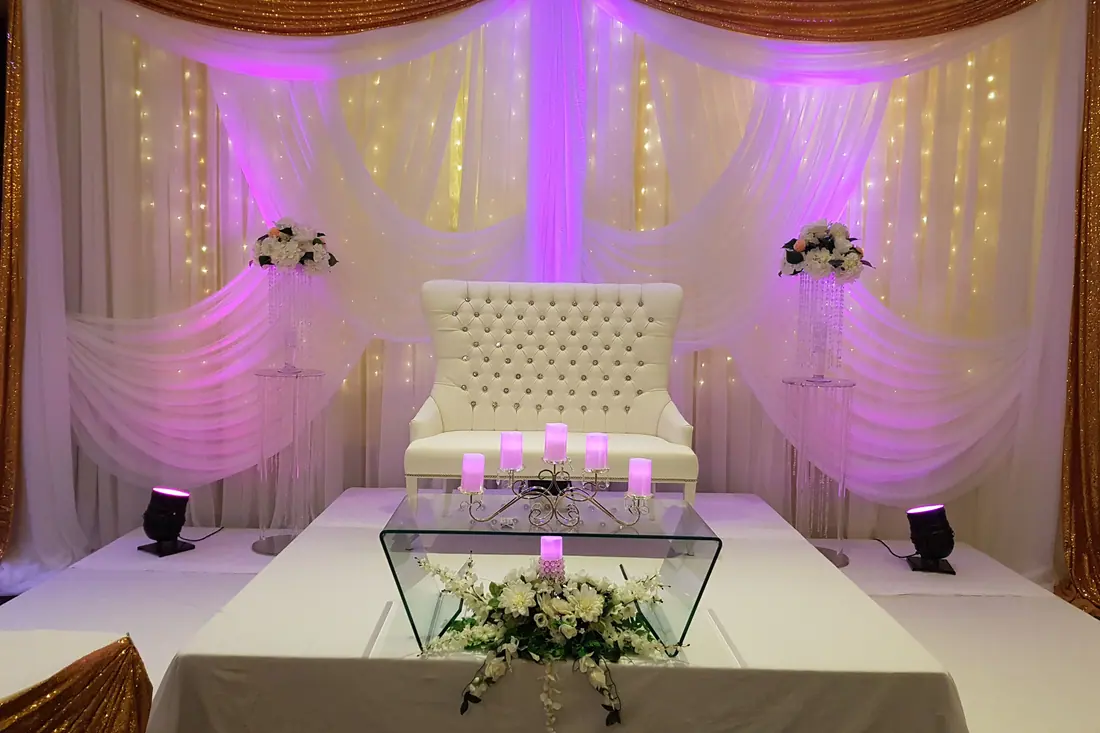Gold sequin and ivory draped backdrop with swagged fabric, a tufted loveseat and a glass sweetheart table of white roses