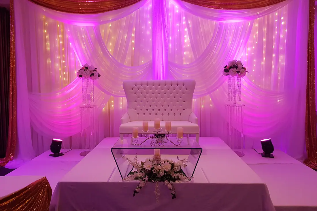 Swagged ivory and gold wedding backdrop washed in magenta uplighting with tall floral pedestals either side of a tufted loveseat