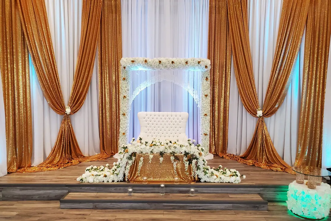 Floor-to-ceiling gold sequin wedding backdrop with pinched ivory draping behind a white tufted throne and gold table