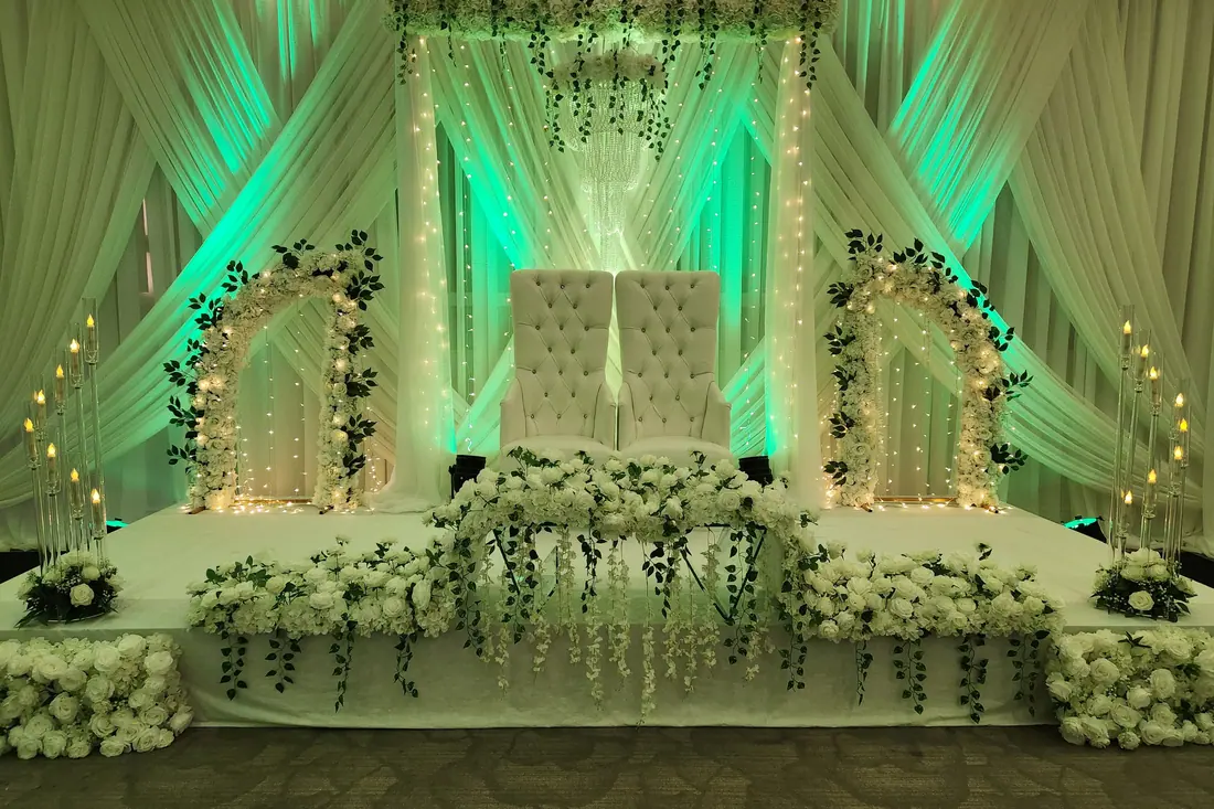 Wedding stage framed by greenery-wrapped arches and fairy lights in soft green uplighting with white florals along the edge