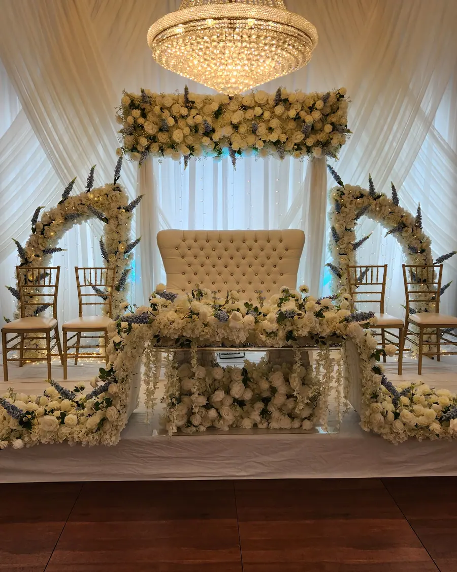 Ivory and gold wedding stage with twin floral arches, a tufted loveseat and a mirrored sweetheart table beneath a crystal chandelier