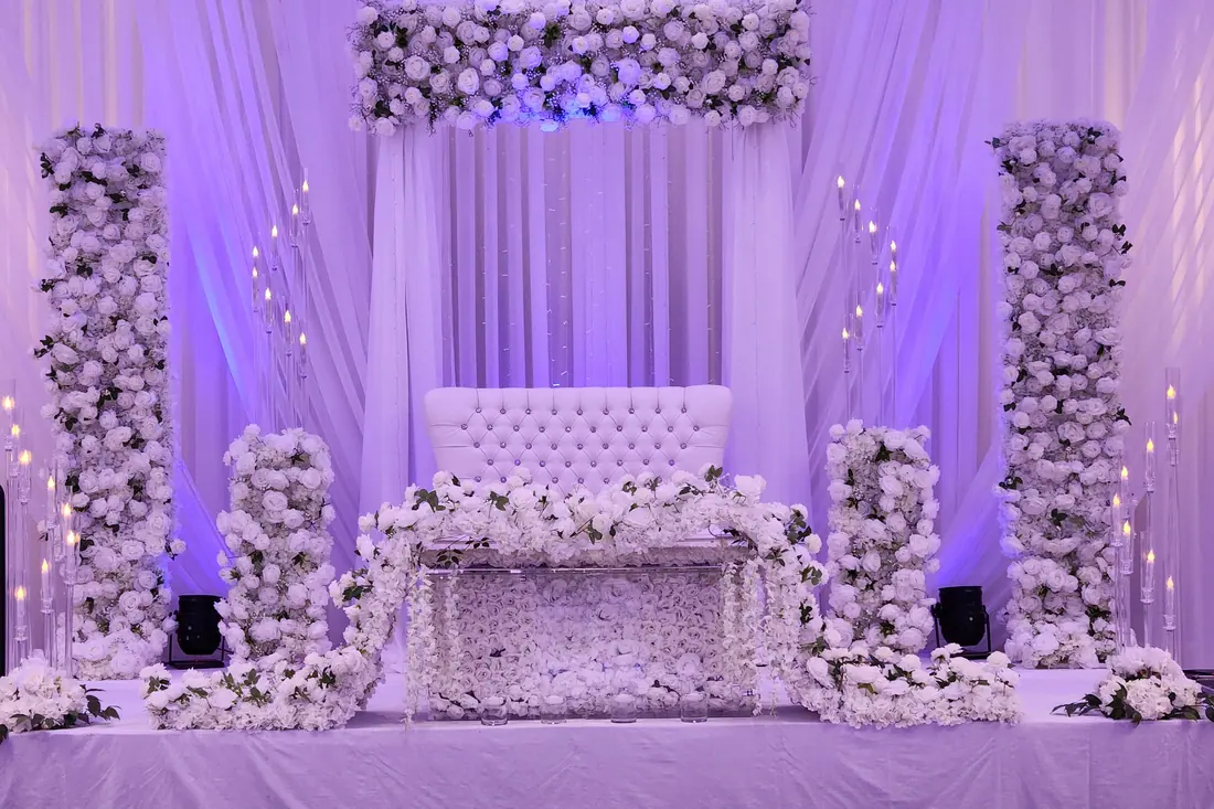 Ivory draped wedding backdrop with lavender floral columns and a white floral valance under violet uplighting