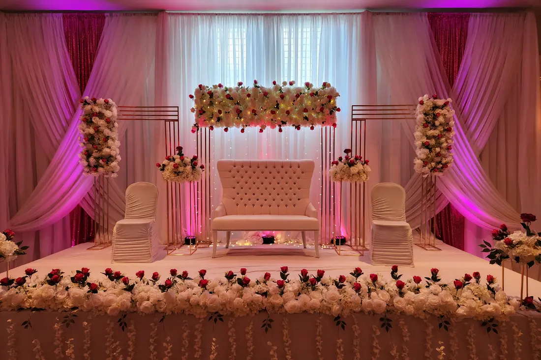 Blush floral wall panel and hanging floral columns lit in warm magenta behind a pale tufted wedding bench