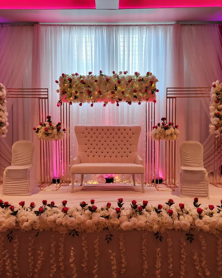 Blush floral wall panel and hanging floral columns lit in warm magenta behind a pale tufted wedding bench