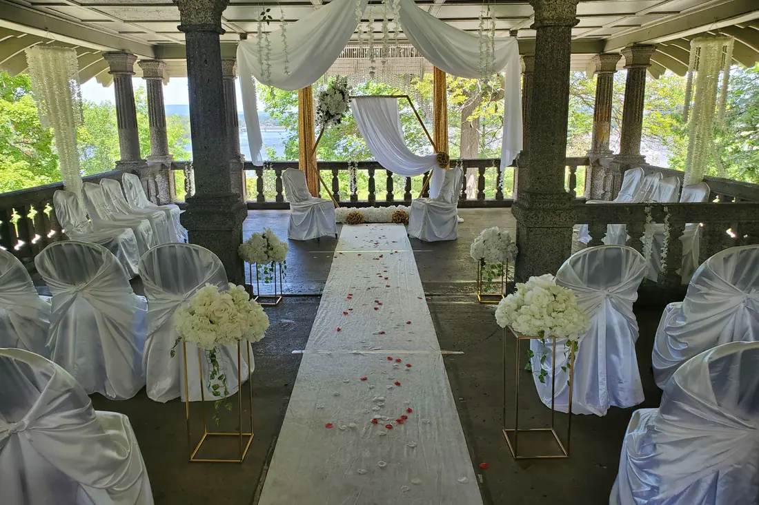 Outdoor wedding ceremony in a stone gazebo with white draping, petal-strewn aisle runner and white covered chairs tied with organza