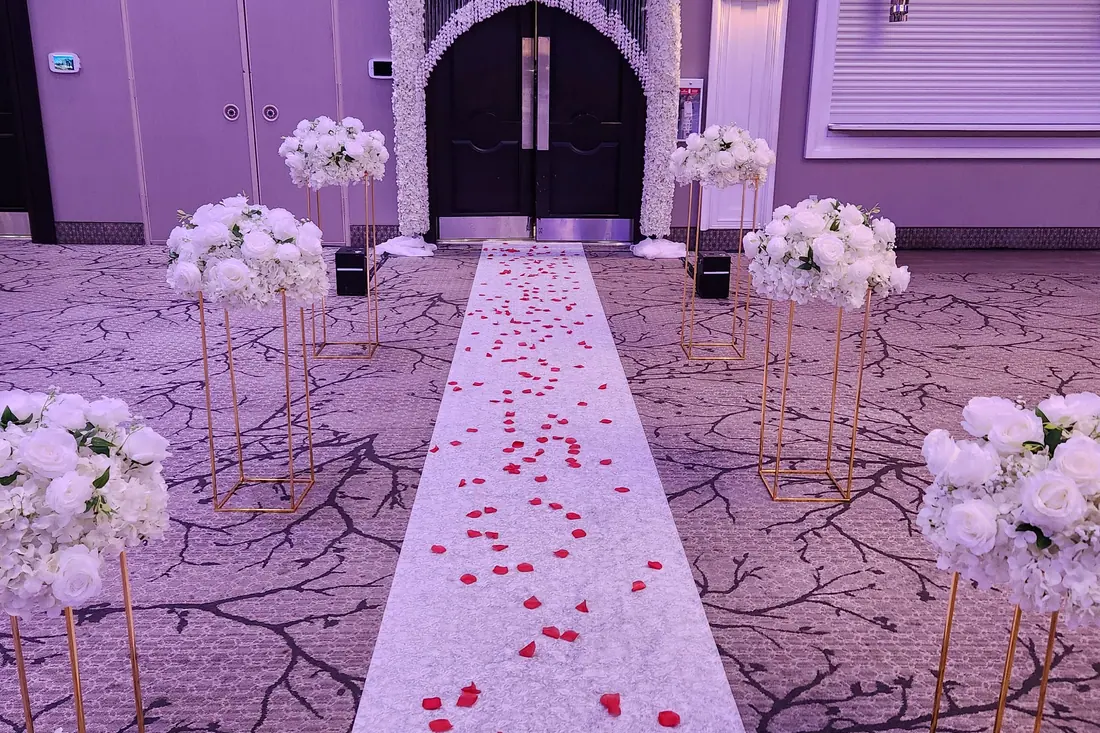 White aisle runner scattered with red rose petals between gold floral stands, lit in soft violet at a ballroom entrance