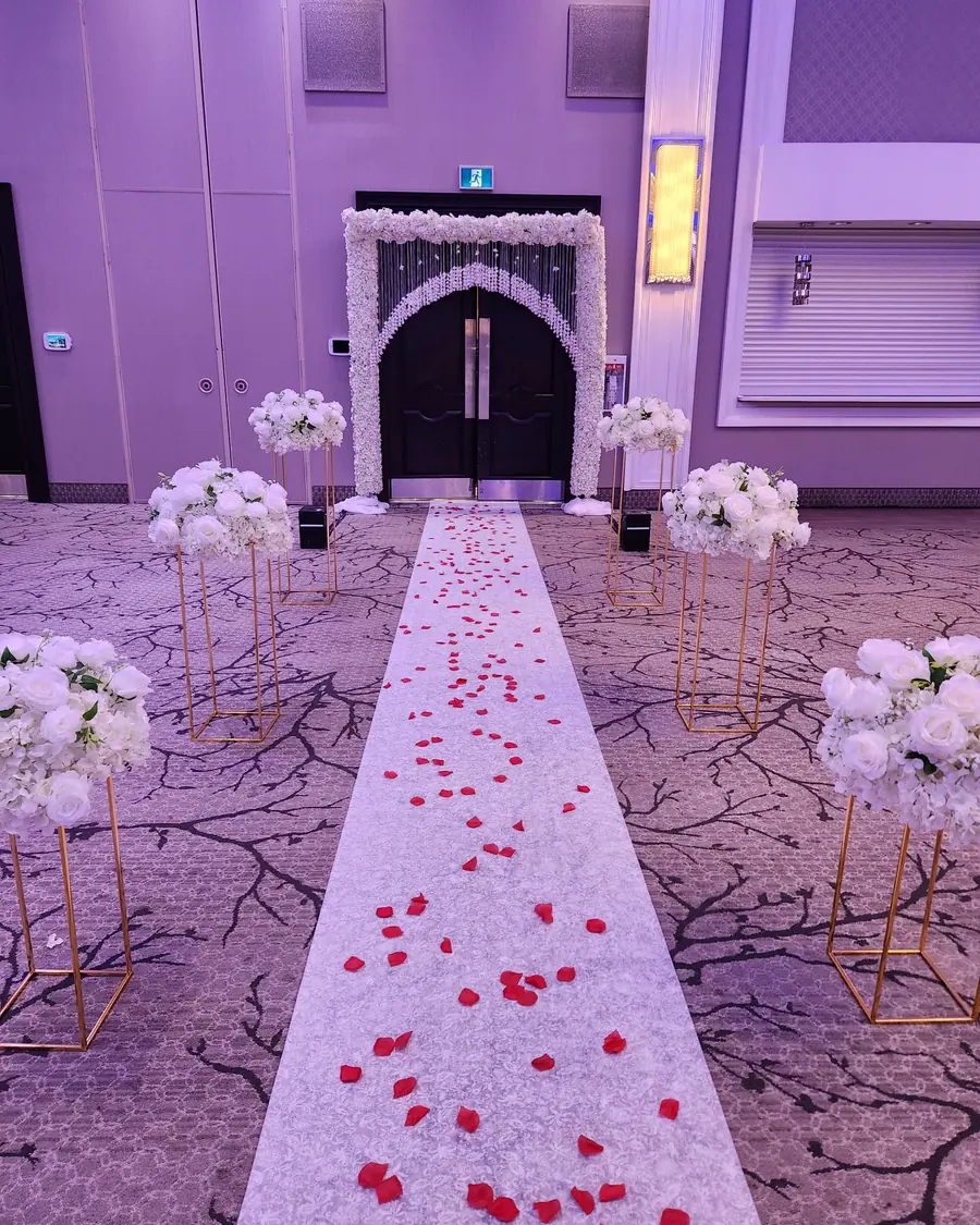 White aisle runner scattered with red rose petals between gold floral stands, lit in soft violet at a ballroom entrance