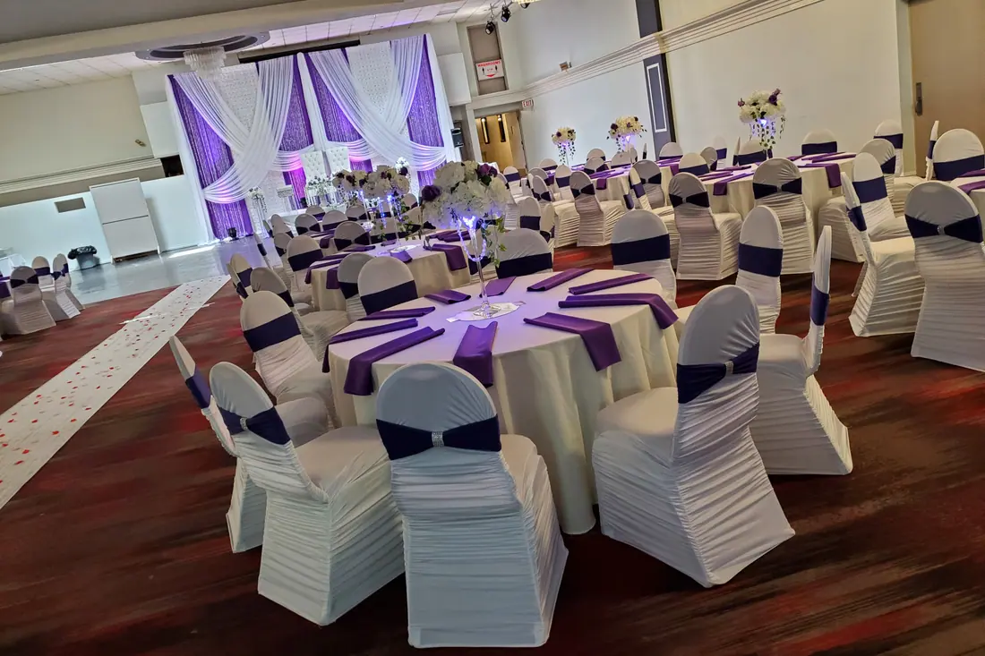 Plum satin overlays layered on white round tables with ruched white chair covers across a hardwood ballroom floor