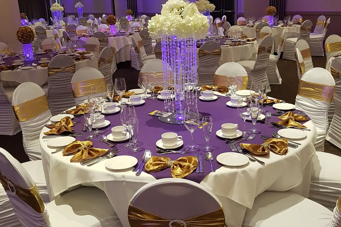 Purple wedding table linen with gold satin napkins and a tall crystal centerpiece crowned with white hydrangea