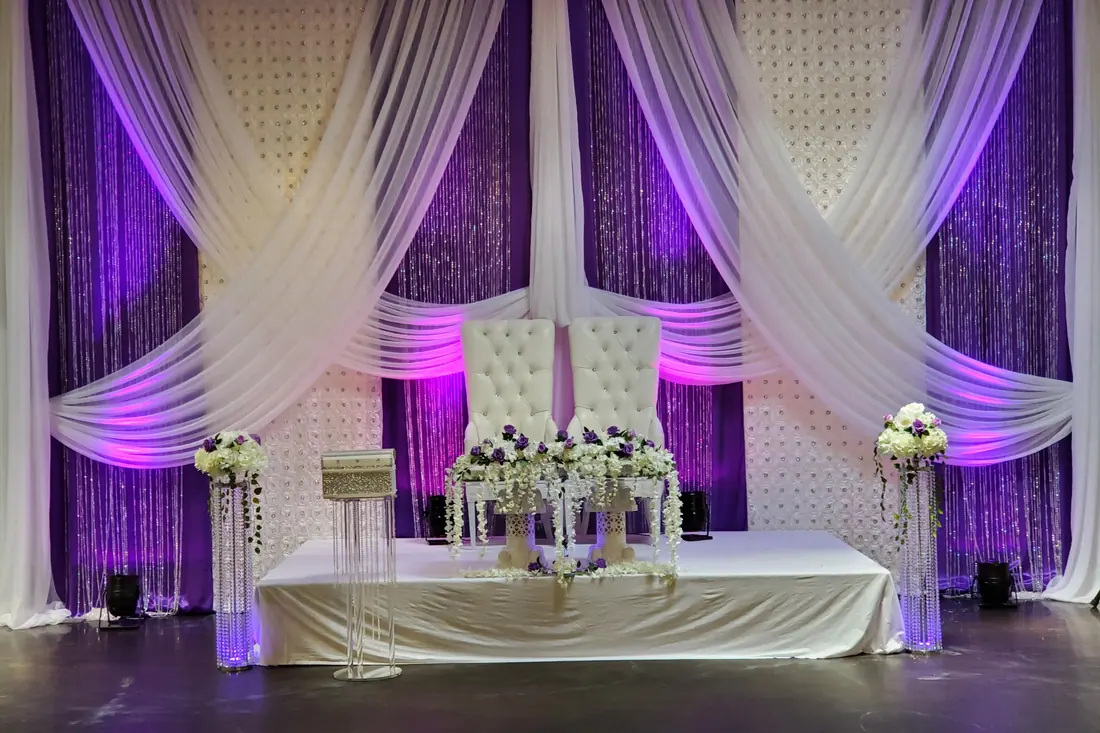 Purple and white sequin wedding backdrop with draped ivory panels behind a tufted throne and crystal pedestal florals