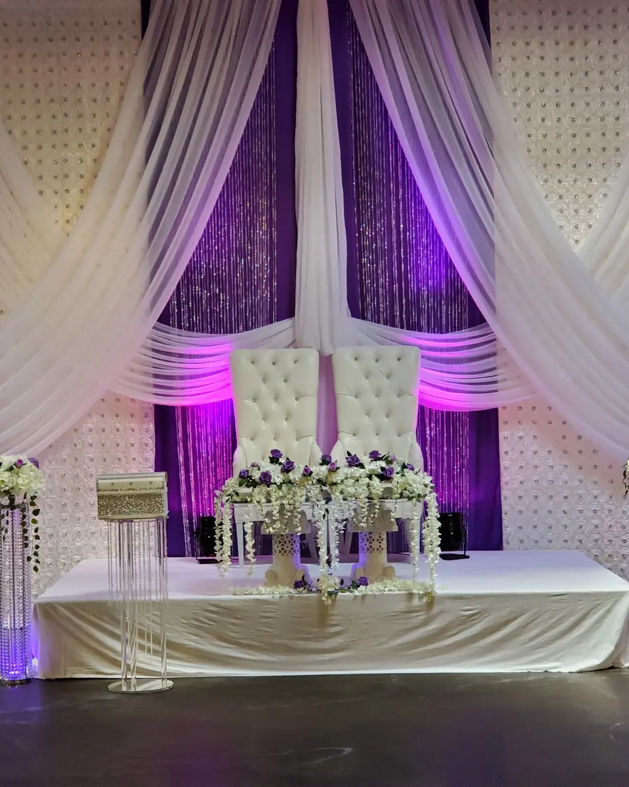 Purple and white sequin wedding backdrop with draped ivory panels behind a tufted throne and crystal pedestal florals