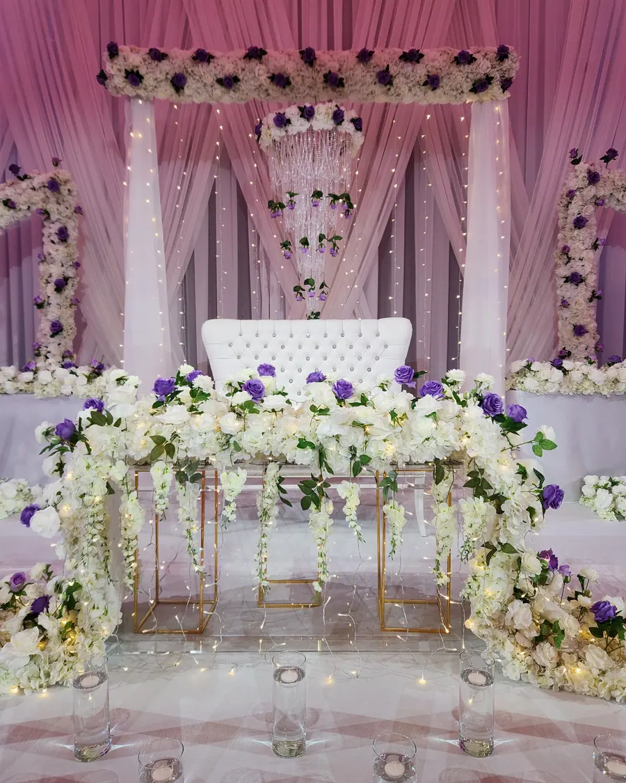 Sweetheart table massed with white and purple blooms and trailing greenery beneath gold floral stands and ivory draping