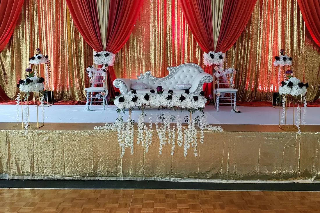 Red and gold pleated wedding backdrop behind a white carved chaise with black and white floral arrangements on gold stands