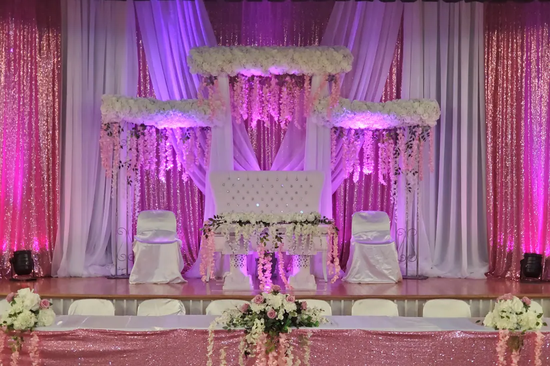 Tiered white floral wedding stage with hanging blossom garlands and ivory draping under pink and violet uplighting