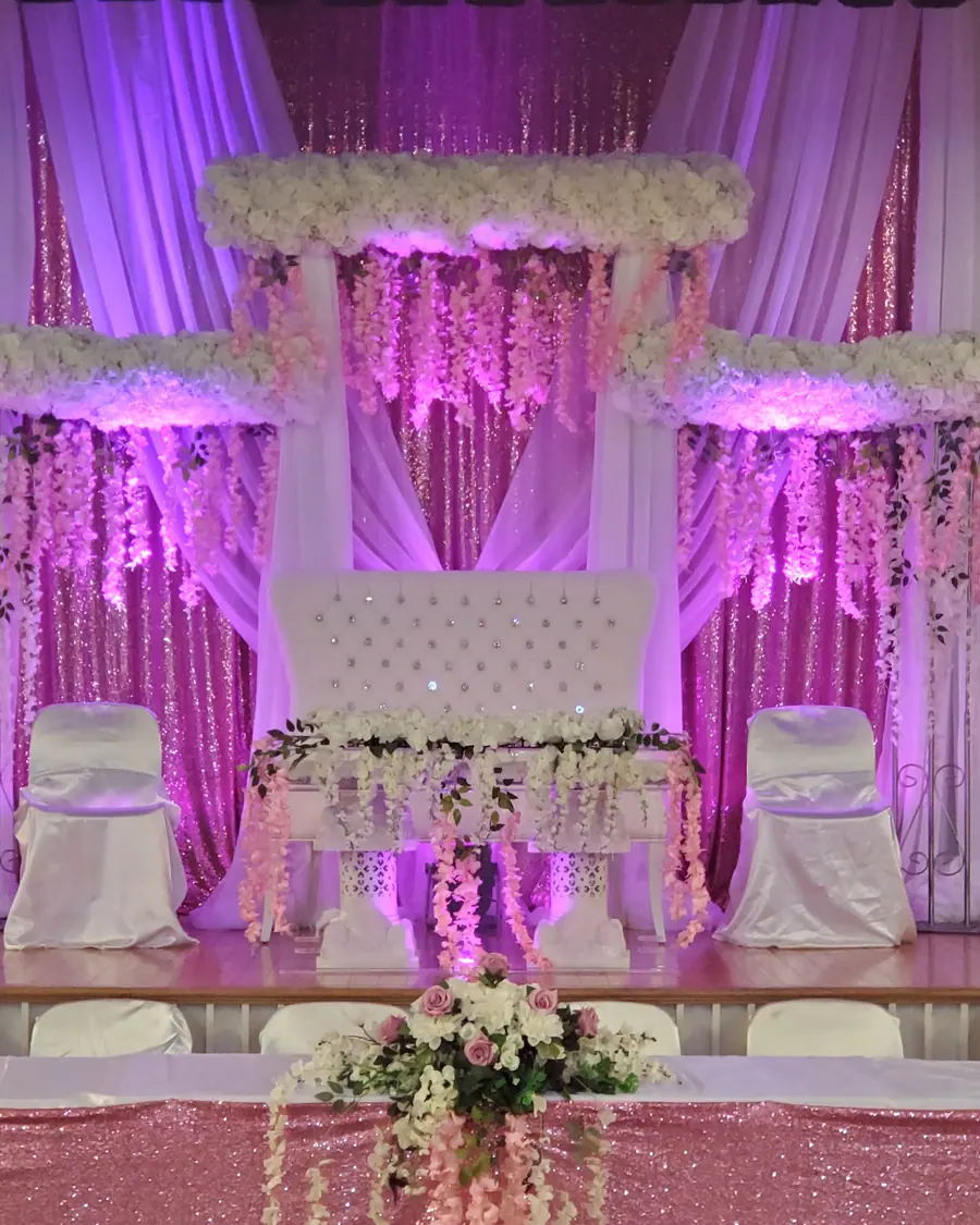 Tiered white floral wedding stage with hanging blossom garlands and ivory draping under pink and violet uplighting