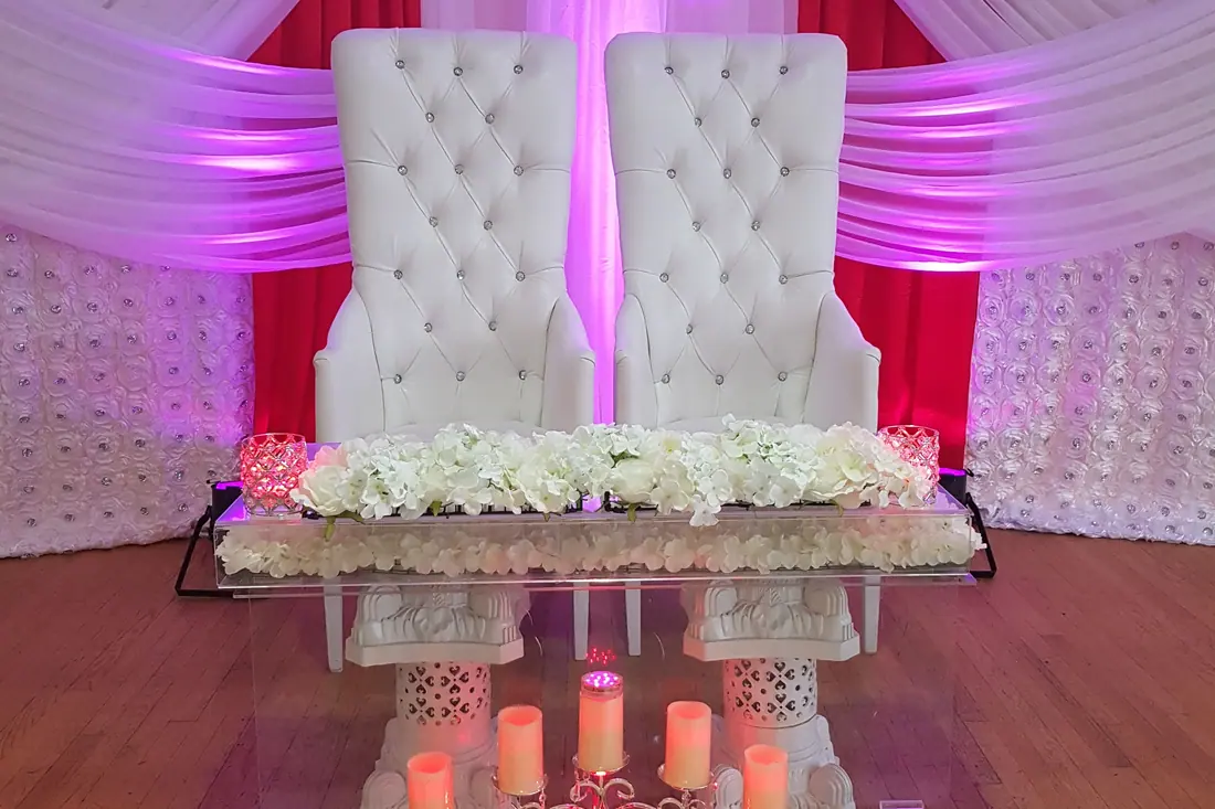 Two tall white tufted throne chairs behind a sweetheart table of white hydrangea, lit in red and fuchsia with pillar candles below