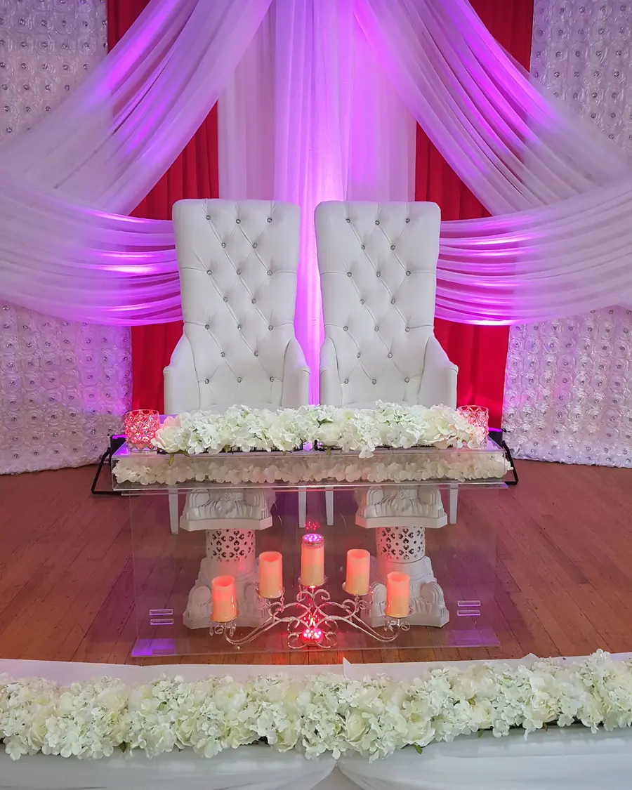 Two tall white tufted throne chairs behind a sweetheart table of white hydrangea, lit in red and fuchsia with pillar candles below