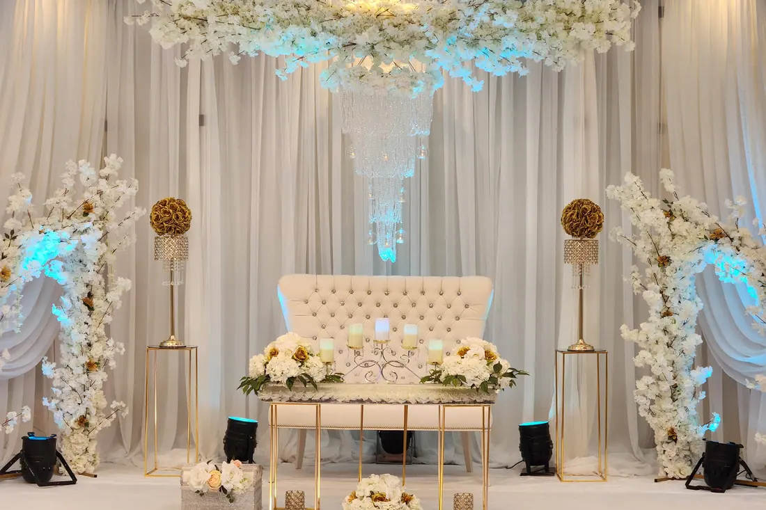 White floral canopy suspended above a tufted loveseat and gold sweetheart table between tall gold floral pedestals